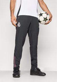 Γκρι αθλητικό παντελόνι με ροζ ρίγες, με λογότυπο στο πάνω μέρος του ποδιού. Το μοντέλο κρατάει μια ασπρόμαυρη ποδοσφαιρική μπάλα.