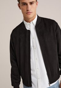 Fekete bomber dzseki sima, matt felülettel, cipzáras záródással és bordázott mandzsettával, egy fehér gombos ingre rétegezve.