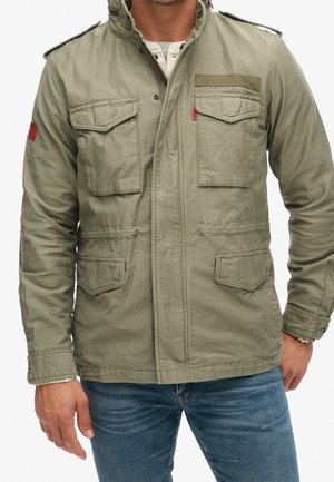 Homme portant une veste utilitaire vert olive avec quatre poches à rabats et un jean bleu, debout avec une main détendue le long du corps.