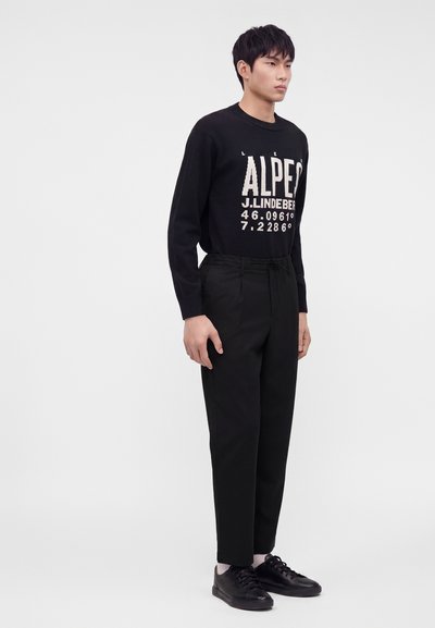 Jeune homme debout portant un pull noir avec un texte, un pantalon noir, des chaussettes blanches et des baskets noires sur un fond clair uni.