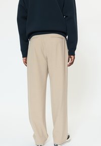 Pantaloni beige su misura con vestibilità a gamba larga, caratterizzati da tasche posteriori e una texture sottile, abbinati a sneakers nere basse.
