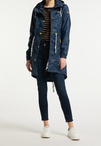 Námořnická voděodolná bunda s kapucí, nastavitelnými šňůrkami, kovovými patentky a dvěma předními kapsami, v kombinaci s tmavými džíny a bílými teniskami.