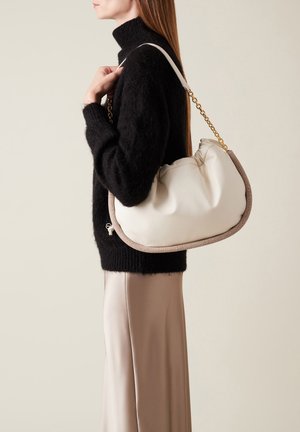 Cream-colored leather handbag with a textured beige trim, a chain strap, and a soft, round design, carried against a black sweater backdrop.