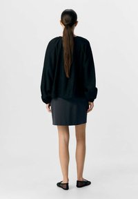Schwarzes strukturiertes Langarmoberteil mit lockerer Passform und einem dunkelgrauen, figurbetonten Rock; das Model steht mit zurückgebundenem Haar und trägt schwarze Mesh-Schuhe.