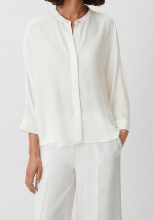 Femme portant un chemisier blanc ample à manches longues avec un col mao et un pantalon blanc large assorti, debout avec une main dans la poche.