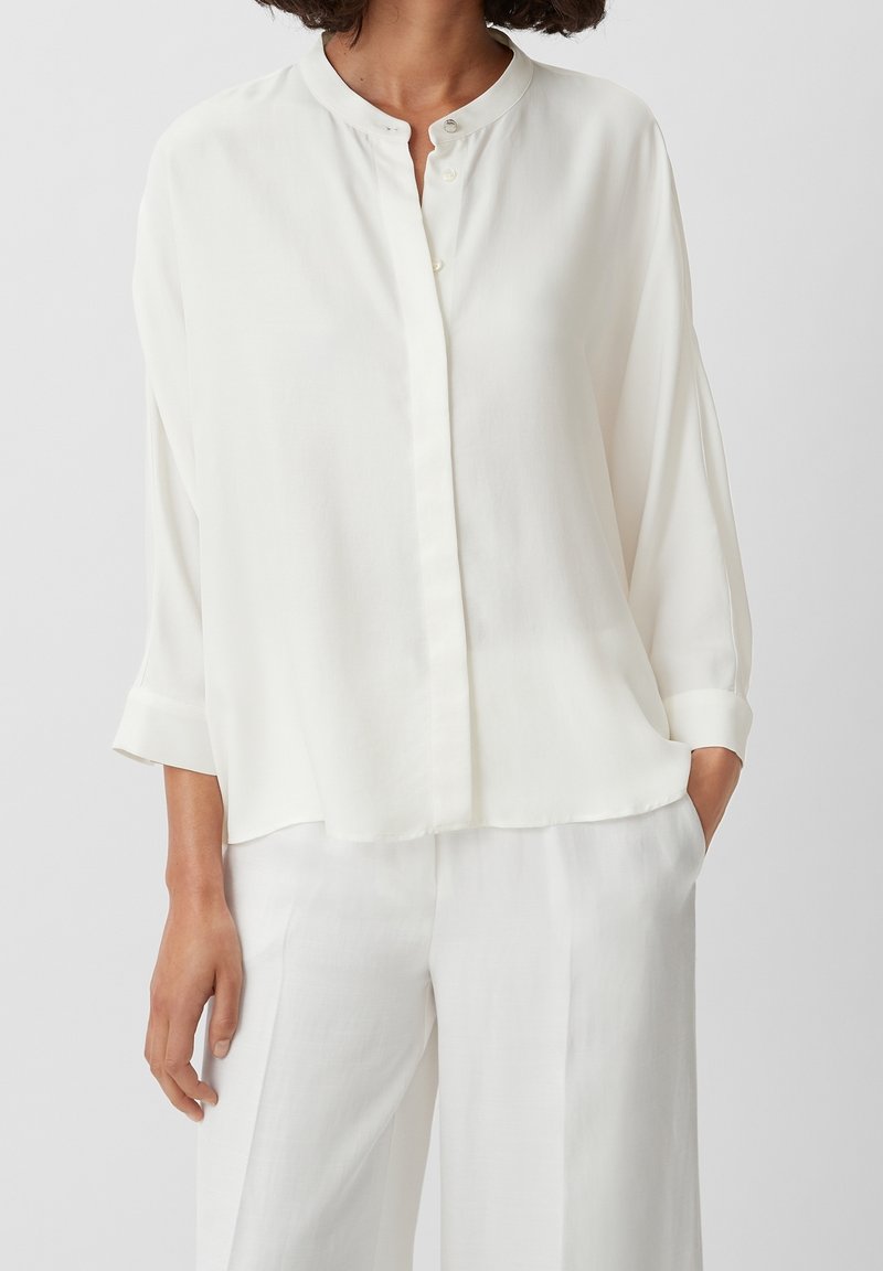 Femme portant un chemisier blanc ample à manches longues avec un col mao et un pantalon blanc large assorti, debout avec une main dans la poche.
