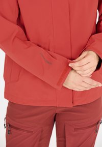Röd jacka med en slät yta, som har justerbara kardborrefästen och en logotyp, tillsammans med matchande röda byxor och dragkedjefickor.