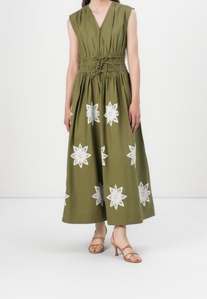 Długa sukienka bez rękawów w oliwkowej zieleni z dekoltem w serek, zaciśnięta w talii za pomocą wiązań, ozdobiona białymi kwiatowymi wzorami, noszona z beżowymi sandałami na obcasie z paskami.
