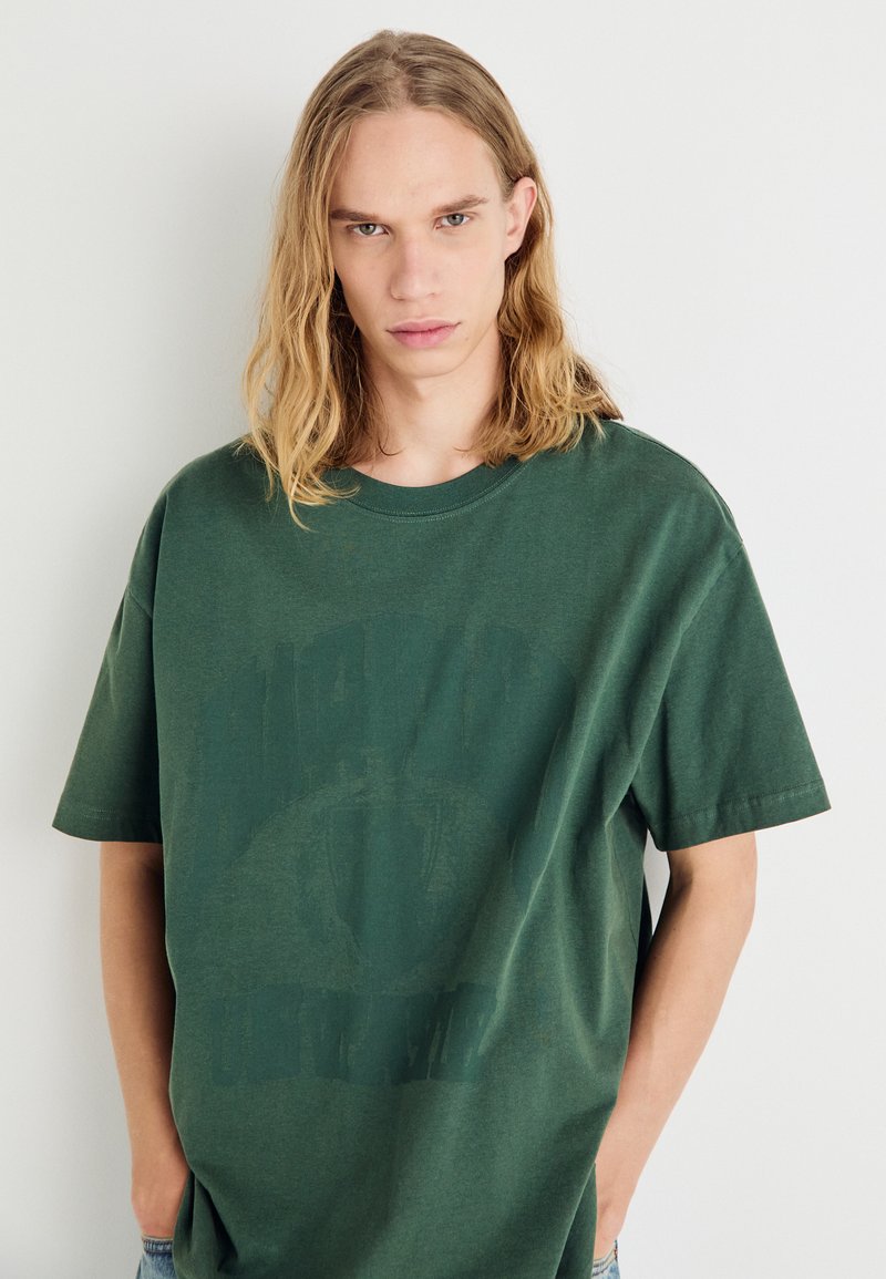 Camiseta verde de gran tamaño, con cuello redondo y hecha de algodón, que presenta un estampado gráfico descolorido en la parte delantera. Manga corta y corte holgado.