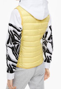 Žlutá nafouknutá vesta s prošívanou texturou, kterou doplňuje bílá a černá mikina s dlouhým rukávem se zebrovaným vzorem. Viditelné šedé tepláky.