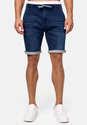 Pantalones cortos de mezclilla en azul oscuro, con dobladillo enrollado, cinturilla elástica con cordón ajustable y dos bolsillos frontales. Combinados con zapatillas blancas.