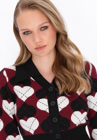 Pletený kardigán s bordovou, čiernou a bielou srdcovou potlačou. Má čierny golier, zapínanie na gombíky a jemnú textúru.