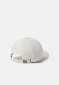 Beige baseball cap with a curved brim, textured fabric, adjustable strap at the back, and embroidered logo detail on the rear.
