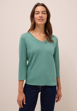Femme portant un t-shirt à manches longues en V turquoise et un jean bleu foncé se tenant devant un fond beige uni.