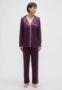 Vijolična pižama iz mehkega blaga. Vključuje srajco z gumbi, okrašeno z belim trakom in žepom, ter hlače v enakem slogu.
