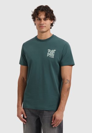 Man met krullend haar in een donkergroene "PURE PATH" logo t-shirt en grijze jeans, staande in een ontspannen houding tegen een effen achtergrond.