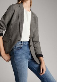 Harmaa houndstooth-tyylinen bleiseri mustilla hihoilla, yksipainikekiinnitys, muotonsa mukainen istuvuus, valkoisen topin päällä ja yhdistettynä sinisiin farkkuihin.