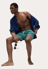 Granatowa koszula z krótkim rękawem, rozpięta, na tealam! kąpielówkach. Model trzyma czarne okulary przeciwsłoneczne, siedzi z odkrytymi stopami. Gładkie faktury i luźny krój.