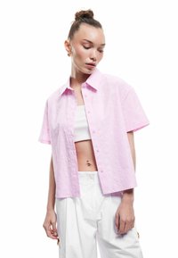 Kortärmad rosa skjorta med texturerat blommönster, knäppning och avslappnad passform, parat med en vit kort topp och vita byxor.