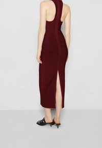 Burgundské přiléhavé šaty s vysokým výstřihem a bočním rozparkem, se žebrovanou texturou a minimalistickým designem. Nosené s černými lodičkami s otevřenou špičkou.