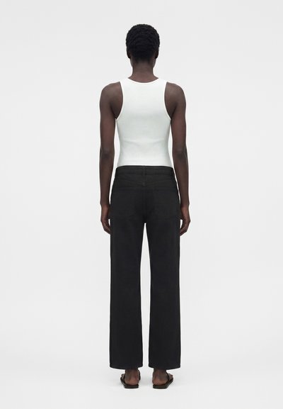 Personne debout de dos portant un haut blanc sans manches côtelé, un pantalon noir à jambes larges et des sandales noires sur un fond uni.