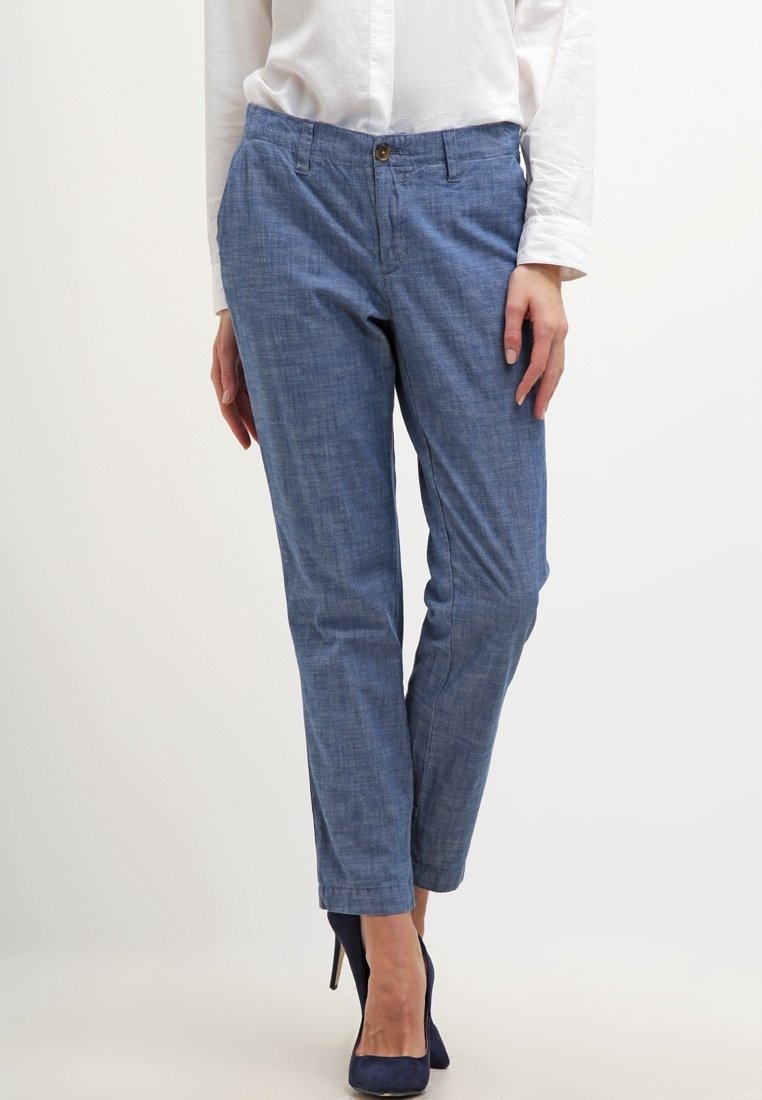Femme portant un pantalon slim bleu, une chemise blanche à manches longues et des chaussures à talons hauts bleu marine, debout devant un fond uni.