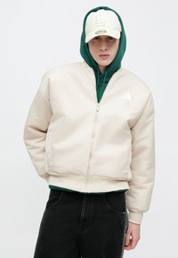 Beež vattitud bomber-tüüpi jakk soonikkoes ääre, tõmbluku ja valge logoga, kantud rohelise kapuutsiga pusa ja mustade teksade peal.