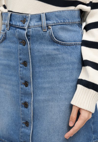Γυναίκα που φοράει μπλε τζιν φούστα με κουμπιά και κρεμ πουλόβερ με μαύρες ρίγες, με το χέρι να ακουμπά στο πλάι, εμφανιζόμενη από τη μέση έως το μέσο του μηρού.