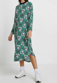 Grön klänning med långa ärmar, som har en svart krage och ett tryck av tigeransikten på en randig bakgrund. Midjekort med sidoklyftor.