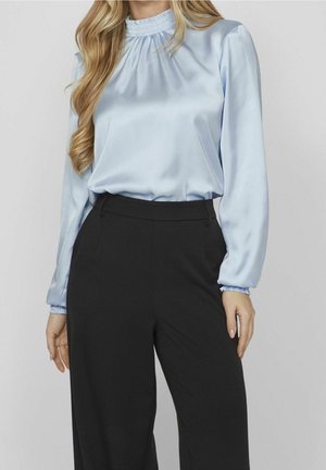 Femme portant un chemisier en satin bleu clair à manches longues rentré dans un pantalon noir taille haute, debout devant un fond uni.