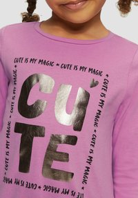 Paarse langekleurige shirt met lange mouwen, voorzien van een grote metallic zilveren "CUTE" grafiek met herhaaldelijke tekst en hartdetails eromheen, van een soepele katoenen textuur.