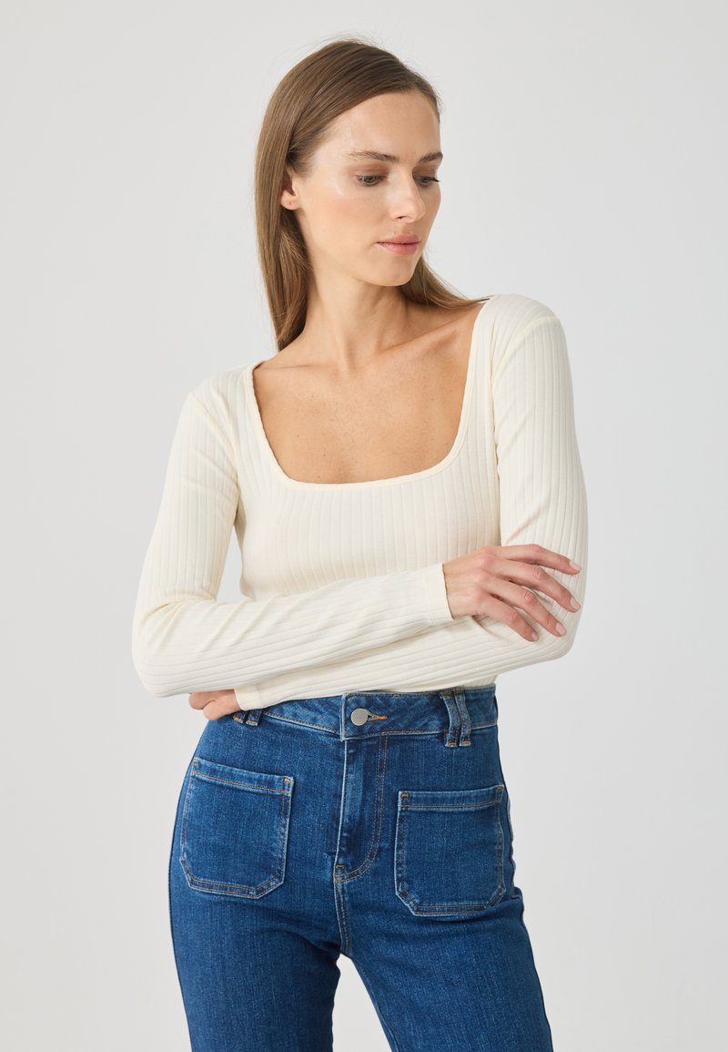 Cămașă cu mâneci lungi, de culoare crem, cu decolteu pătrat, asociată cu blugi din denim de culoare albastru închis, cu talie înaltă și buzunare pe spate.