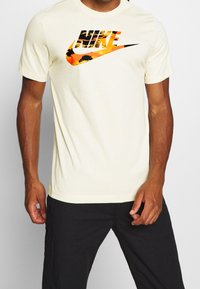 Krämfärgad bomullströja med en djärv Nike-logotyp i orange och svart, med en texturerad grafisk design på framsidan. Korta ärmar.