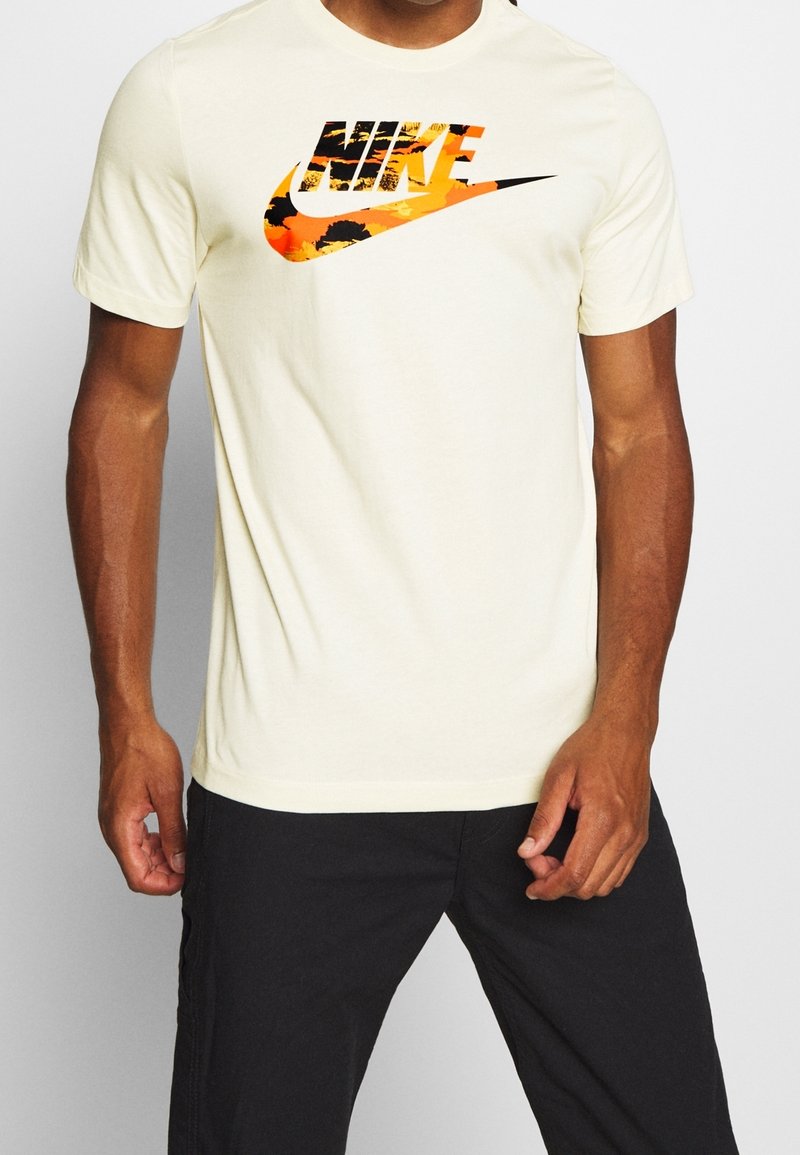 Krämfärgad bomullströja med en djärv Nike-logotyp i orange och svart, med en texturerad grafisk design på framsidan. Korta ärmar.