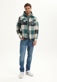 Flanellen shirt in teal, grijs en crème geblokt patroon; twee borstzakken; knoopsluiting aan de voorkant; gelaagd over een grijze hoodie; gecombineerd met blauwe spijkerbroeken en witte sneakers.