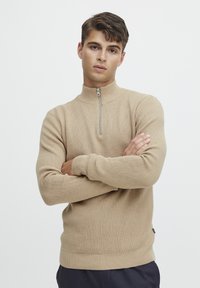 Beige strikkegenser med halvdreppekrage, ribbede mansjetter og nederkant, med et teksturert mønster og en liten merkevaredetalj på siden.