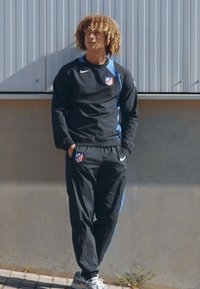 Black athletic tracksuit with blue accents, featuring a team logo, and grey sneakers. The outfit has a sporty design and casual fit.