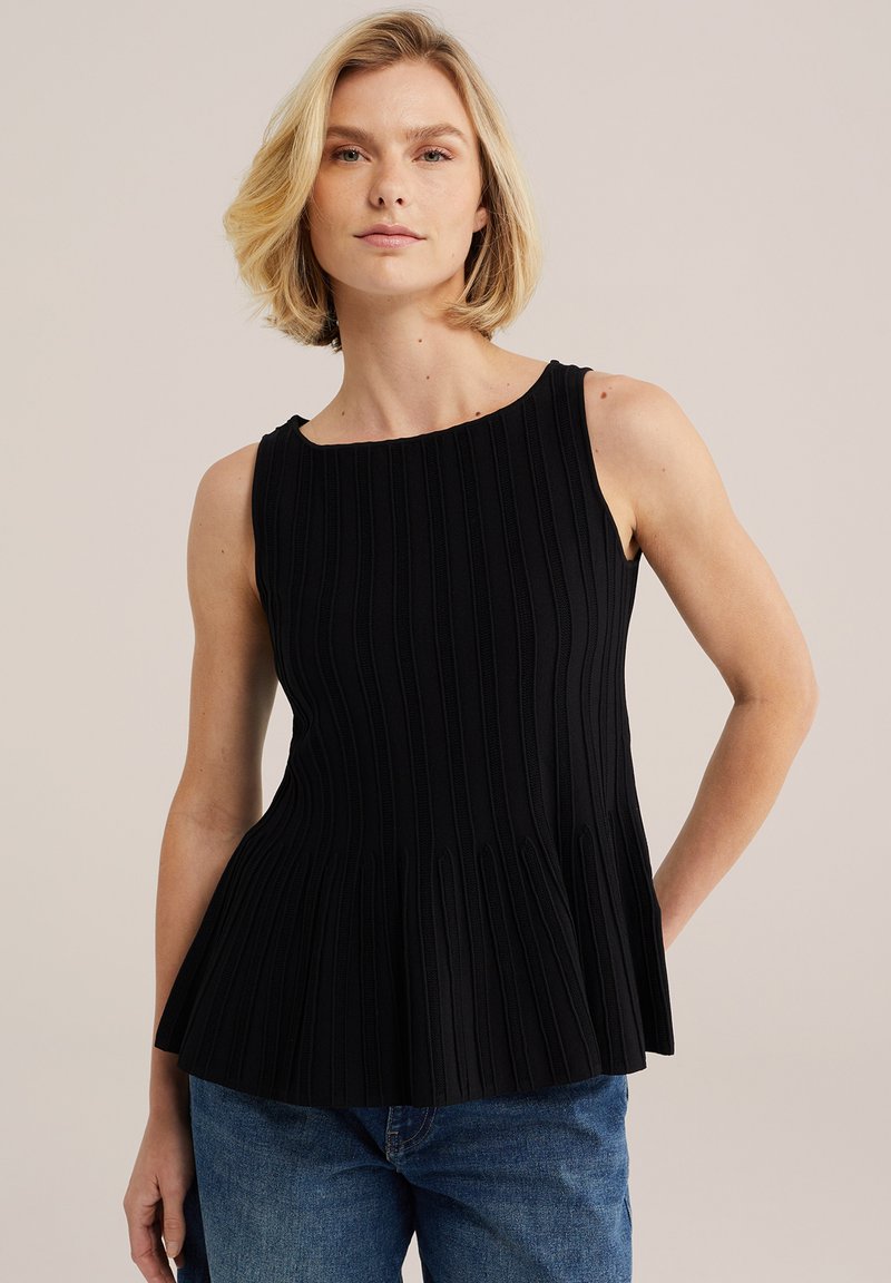 Femme blonde portant un haut noir sans manches en maille côtelée avec un ourlet évasé et un jean bleu, debout devant un fond beige clair.