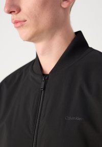 Black bomber jacket with a zip closure, ribbed collar, and a subtle logo on the chest. Smooth, lightweight fabric with a matte finish.