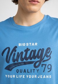 Mėlyna medvilninė marškinėliai su dideliu, tamsiai mėlynu vintage stiliaus tekstu. Tekste yra "BIG STAR", "QUALITY '79" ir "YOUR LIFE YOUR JEANS."
