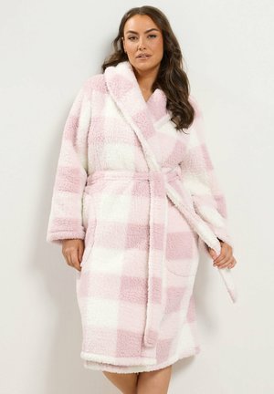 Femme portant un peignoir polaire à carreaux roses et blancs jusqu’au genou, noué à la taille, debout devant un fond clair uni.