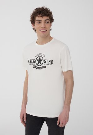 T-shirt bianco in cotone con un logo a stella stampato in nero e testo. Maniche corte e scollo rotondo. Indossato con jeans scuri.