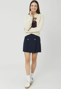 Pull en maille crème avec graphique d'ours, associé à une jupe plissée marine avec des boutons dorés, porté avec des chaussettes blanches et des baskets.