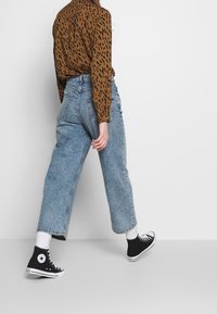 Hnědá košile s zvířecím vzorem, džíny světle modré barvy s vysokým pasem a černé tenisky Converse s bílými podrážkami. Viditelné bílé ponožky.