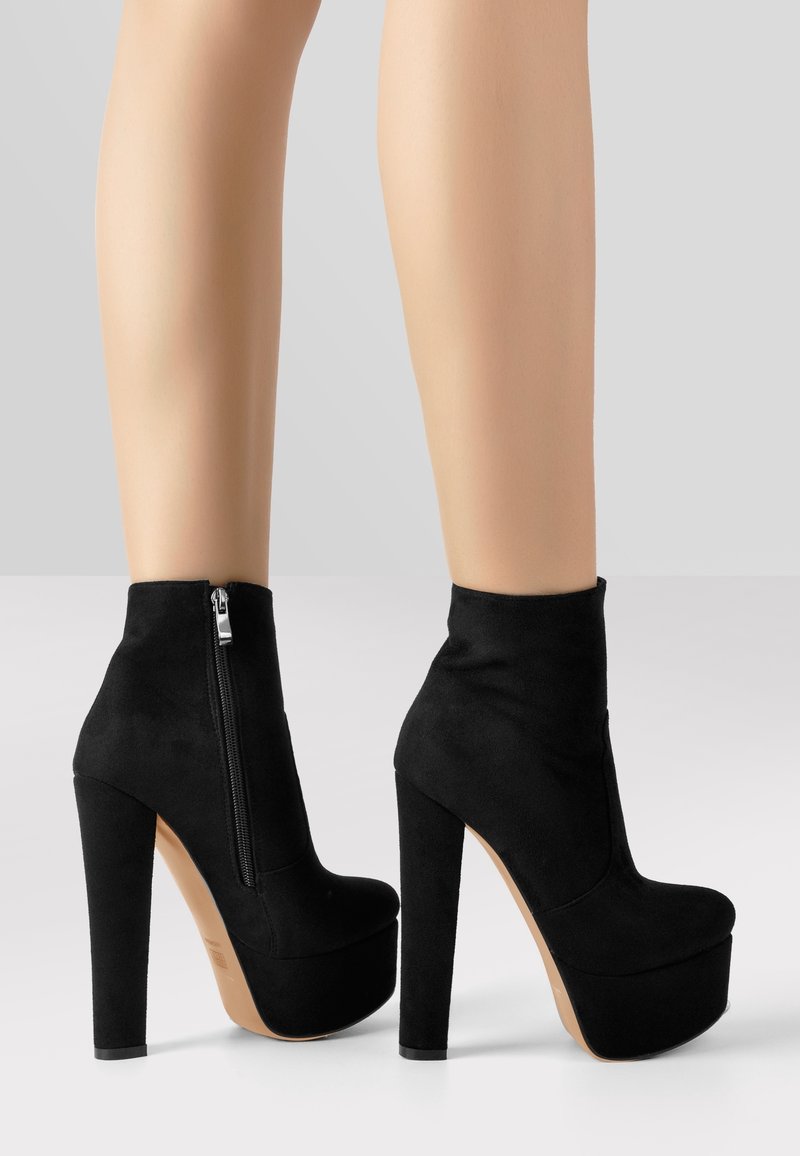 Jambes portant des bottines à semelle plateforme en suède noir, avec talons carrés hauts et fermetures éclair latérales, sur un fond gris clair.