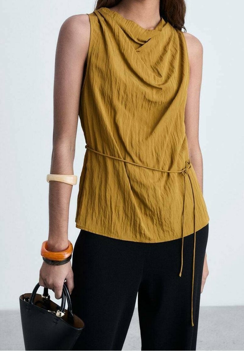 Femme portant un haut sans manches moutarde avec un lien à la taille, un pantalon noir, tenant un sac à main noir et portant des bracelets épais à un bras.