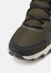 Mørk olivengrønne joggesko med en mesh-overdel og sorte detaljer. Teksturert gummi såle med hvite prikker. Lukkesystemet er synlig.