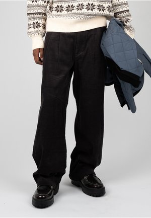 Persoon die zwarte corduroy broek, zwarte loafers, crème trui met sneeuwvlokpatroon draagt en in één hand een blauw doorgestikt jack vasthoudt.