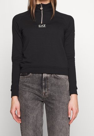 Frau trägt ein schwarzes EA7 Emporio Armani Langarmshirt mit halbem Reißverschluss und hohem Kragen, kombiniert mit ausgewaschenen schwarzen Jeans.
