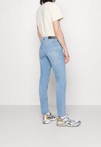 Jeans de mezclilla de color azul claro con un corte ajustado, que cuentan con dos bolsillos traseros y un parche de cuero negro. Combinados con una camiseta beige y zapatillas con detalles en plata.